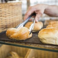 En hand plockar upp en sesamfröbeströdd bulle med tång från en bakplåt i ett bageri.