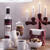 En uppsättning föremål på ett bord inkluderar en flaska, ljusstakar, kaffekoppar, en glasskål med nötter och en burk med kakor under en glaskupa.