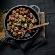 En gjutjärnsgryta fylld med köttbullar står på ett träbord med en träslev och en kvist rosmarin bredvid.