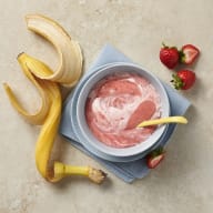 En skål med rosa kräm står på en servett bredvid en banan och jordgubbar.