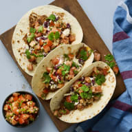 Tre tacos ligger på en skärbräda bredvid en skål med salsa och en vikt handduk.