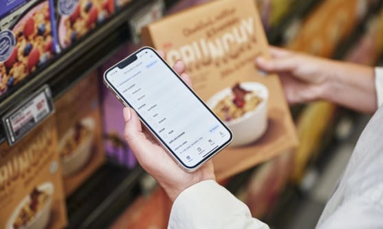 I en butiksmiljö är det en hand som håller i ett paket med flingor och en mobiltelefon med ICA appen och tittar på sin inköpslista.