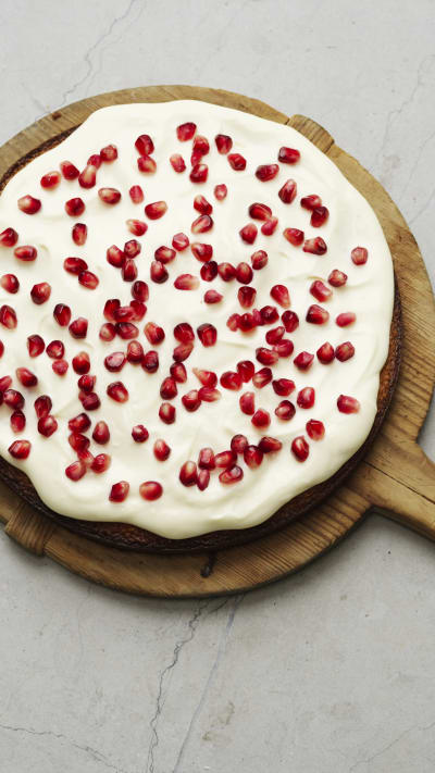 En rund saffranskladdkaka med glasyr och granatäppelkärnor ligger på ett träfat.