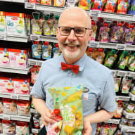 En butikspersonal håller en påse majschips, en fruktpurépåse och en förpackning blöjor framför hyllor med barnmat.