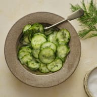 En skål med gurkskivor och dill står på ett bord bredvid en sked och en liten skål.
