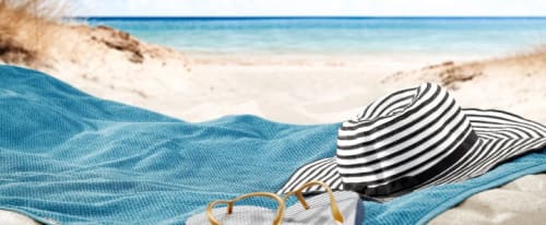 Bild på en handduk, solhatt och ett par tofflor på en sandstrand