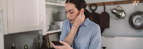 Ung kvinna står i köket med smartphone och sätter i trådlösa hörlurar