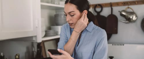 Ung kvinna i köket tittar på mobilen och sätter in trådlösa hörlurar