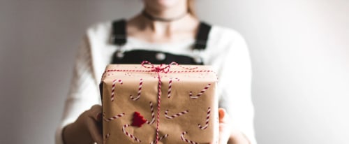 Kvinna i hängselbyxor räcker fram en julklapp med brunt papper och polkagrismönster.
