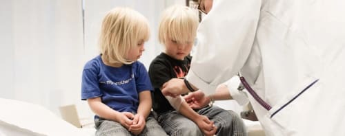 Två barn som sitter på en säng och en läkare som står bredvid.