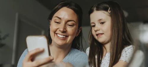 Mamma och dotter som sitter vid ett köksbord och tittar på en mobiltelefon tillsammans.