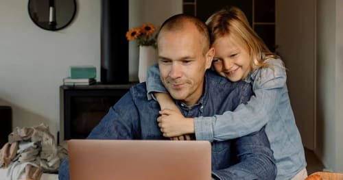 Pappa blir kramad av sin dotter när han sitter framför en bärbar dator.