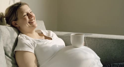En leende gravid kvinna sittligger i en soffa med en tekopp på magen.
