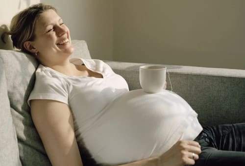 En leende gravid kvinna sittligger i en soffa med en tekopp på magen.