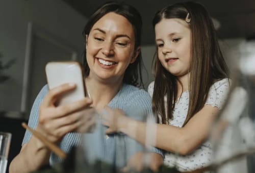 Mamma och dotter som sitter vid ett köksbord och tittar på en mobiltelefon tillsammans.