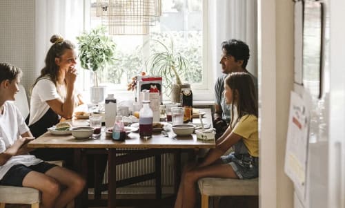 Familj som äter frukost vid ett köksbord framför ett fönster.
