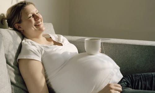 En leende gravid kvinna som liggsitter i en soffa med en tekopp på magen.