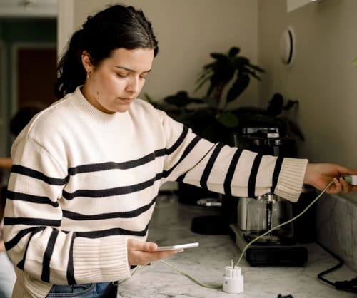 Kvinna laddar sin mobil på en köksbänk. Spartips i vardagen för dig som vill ha mer pengar över i plånboken.