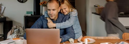 Pappa sitter med laptop vid matbordet och får en kram av sin dotter.