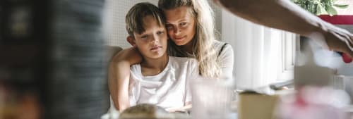 Mamma sitter med sonen i knät vid köksbordet och kramar om honom.