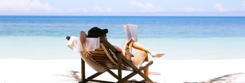 En person i en solstol på en vit strand med turkost hav framför sig