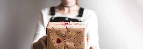Kvinna i hängselbyxor räcker fram en julklapp med brunt papper och polkagrismönster.