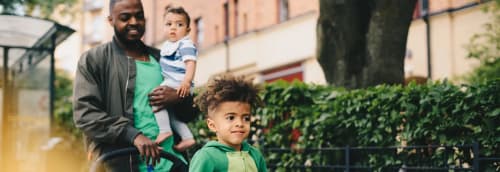 Man promenerar med en baby på armen och ett småbarn i vagnen.