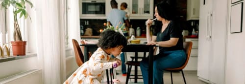 En småbarnsfamilj i köket. Föräldrarna gör vardagssysslor medan ett barn leker med sparkcykel.