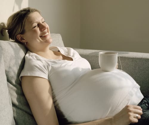 Gravid kvinna som ligger i en soffa med en tekopp på magen.
