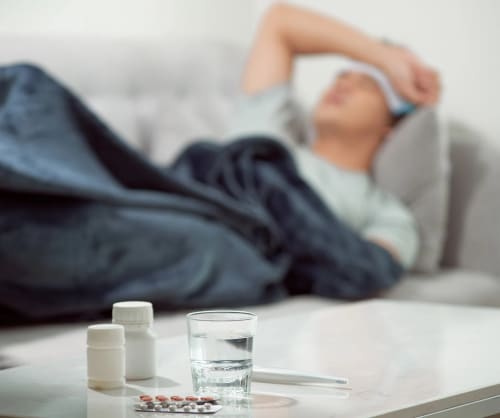 Man som ligger sjuk under ett täcke i soffan med mediciner liggandes på ett bord bredvid.