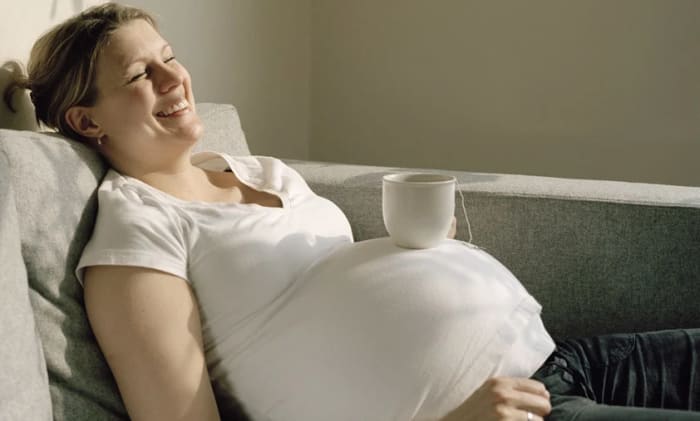 En leende gravid kvinna som liggsitter i en soffa med en tekopp på magen.