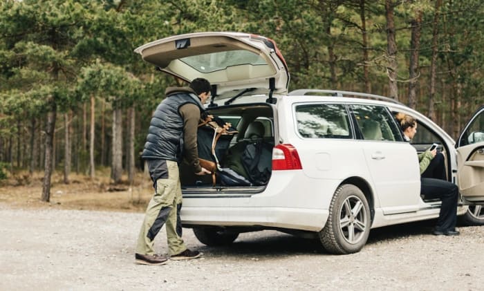 En man packar in en väska i bakluckan på en bil och en kvinna sitter i bilen med öppen dörr.