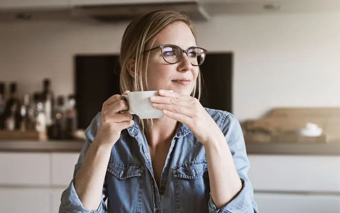 Kvinna som sitter och dricker kaffe och tänker vid ett köksbord tittandes mot ett fönster.