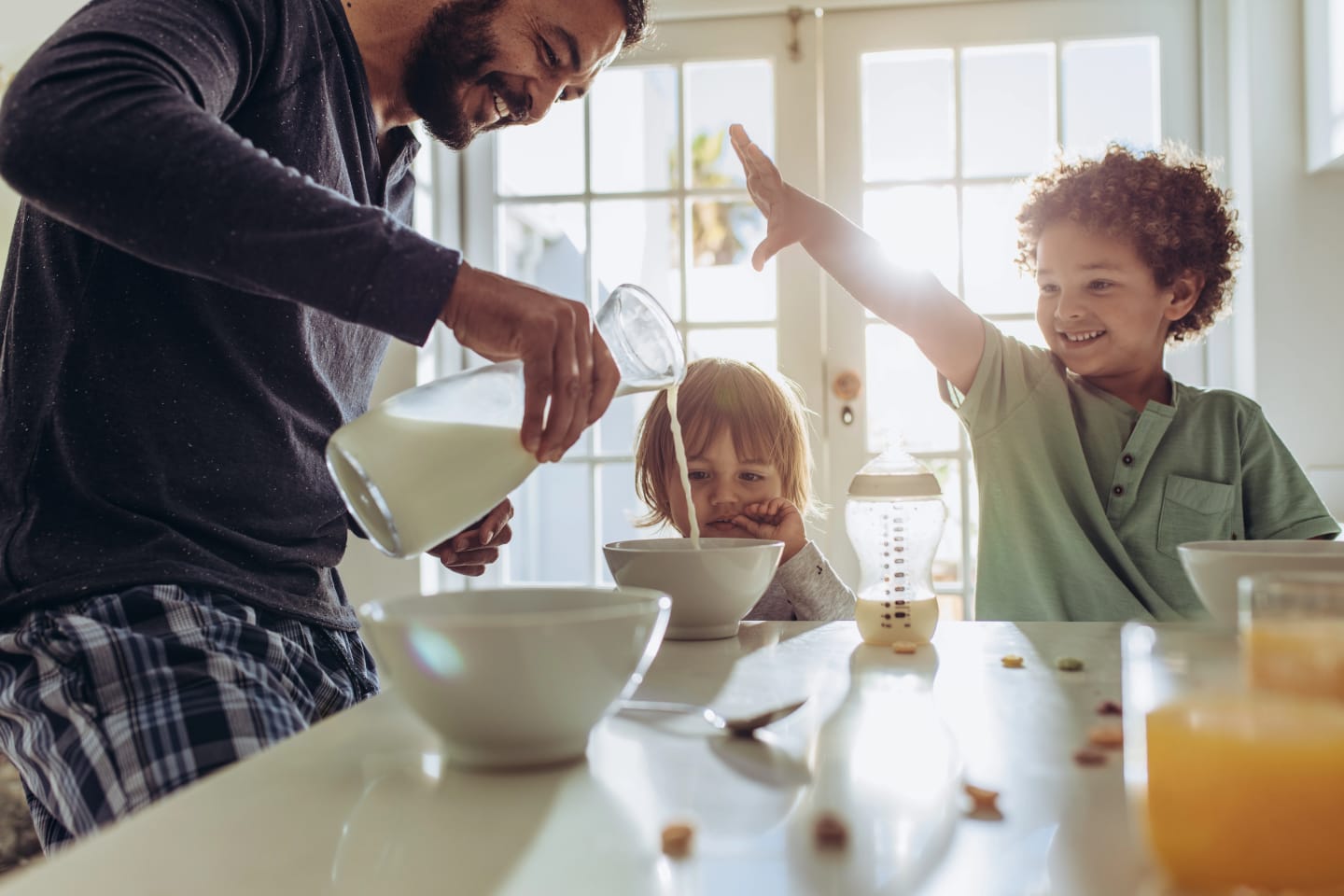 Pappa häller upp mjöl till barn vid frukostbordet. Två barn sitter ner och ler.