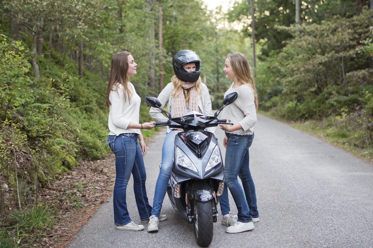 Tre unga tjejer står vid mindre väg. Hon i mitten har mopedhjälm och sitter på sin moped.