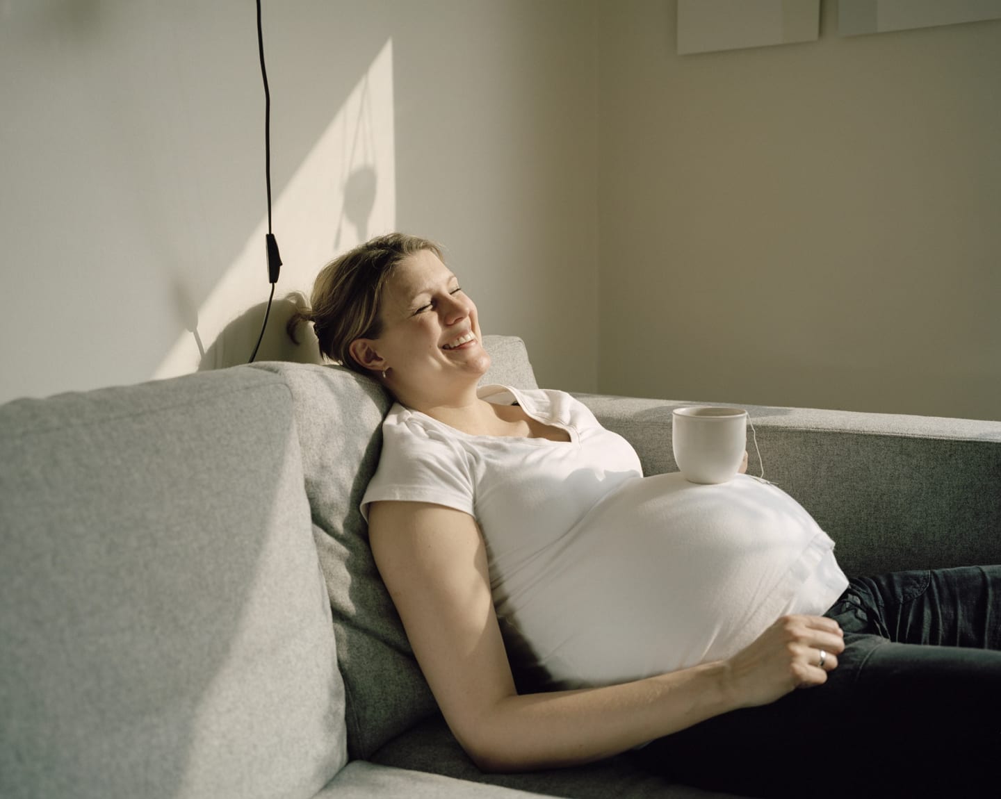 Kvinna ligger i soffan höggravid med en kopp på magen och ler.