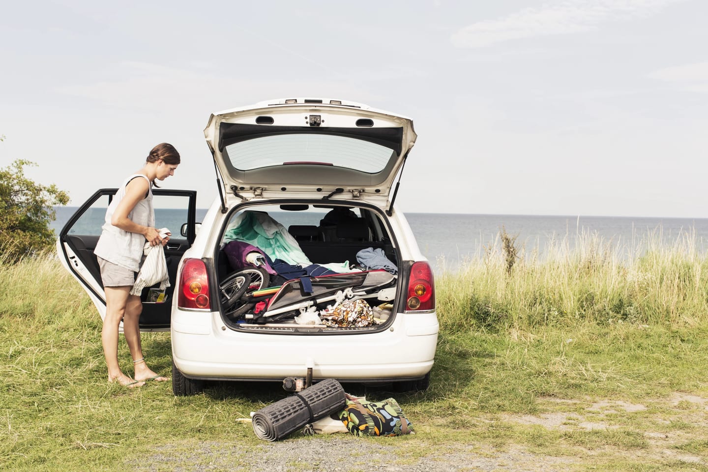 Bil står parkerad bredvid havet med öppen baklucka. Bakluckan är fullpackad och bredvid bilen står en kvinna och tittar in i bilen.