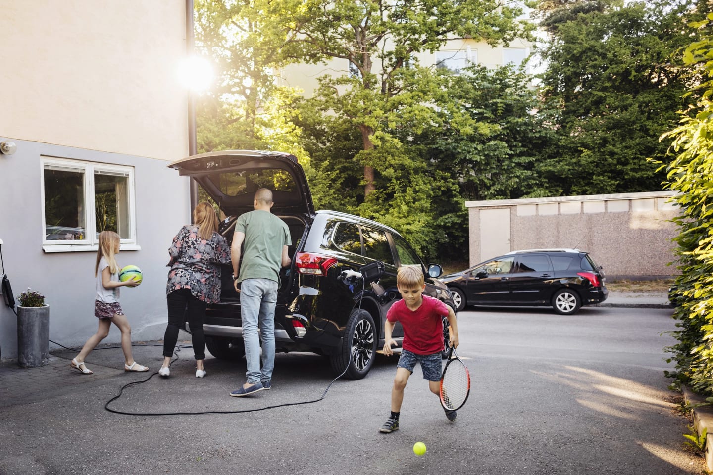 Bil står parkerad med öppen baklucka. Barn leker med tennisracket och boll medan föräldrarna står vid backluckan på bilen och tittar in.