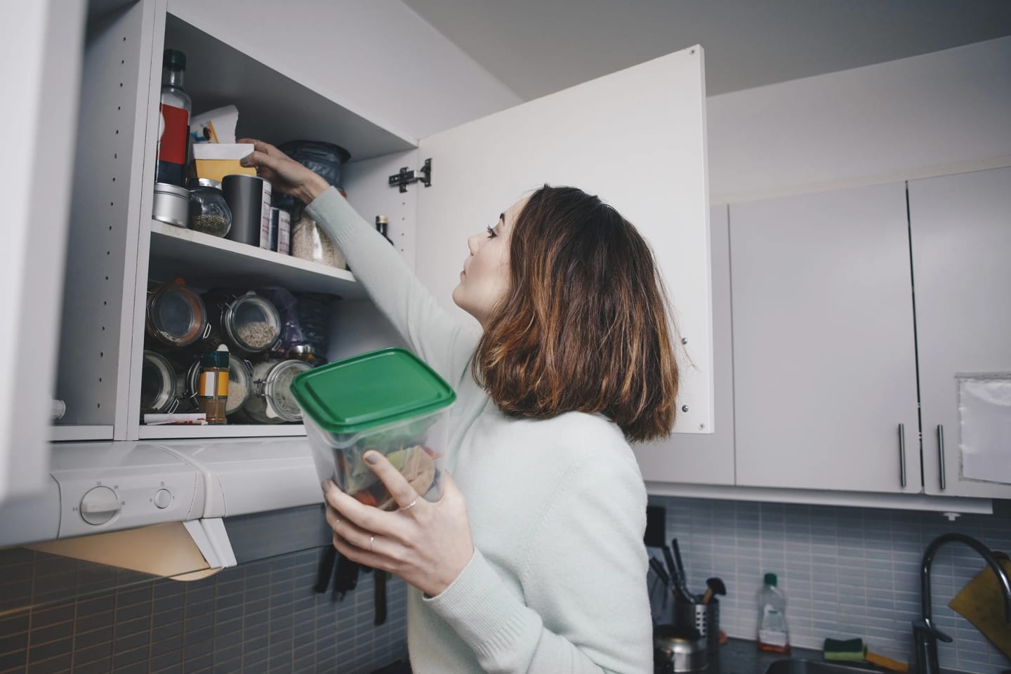 Ung kvinna står i köket med öppet skafferi och sträcker sig för att hämta något. Hon har en plastmatlåda med grönt lock i handen.