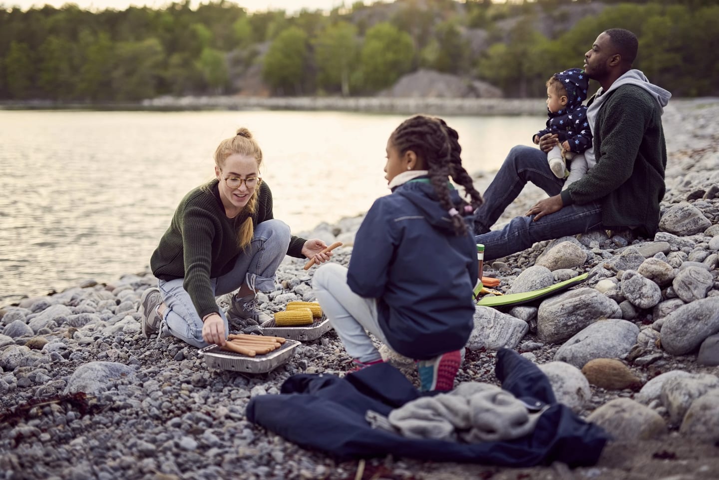 Familj på två barn, en mamma och en pappa som grillar korv vid vattnet på stenstrand.