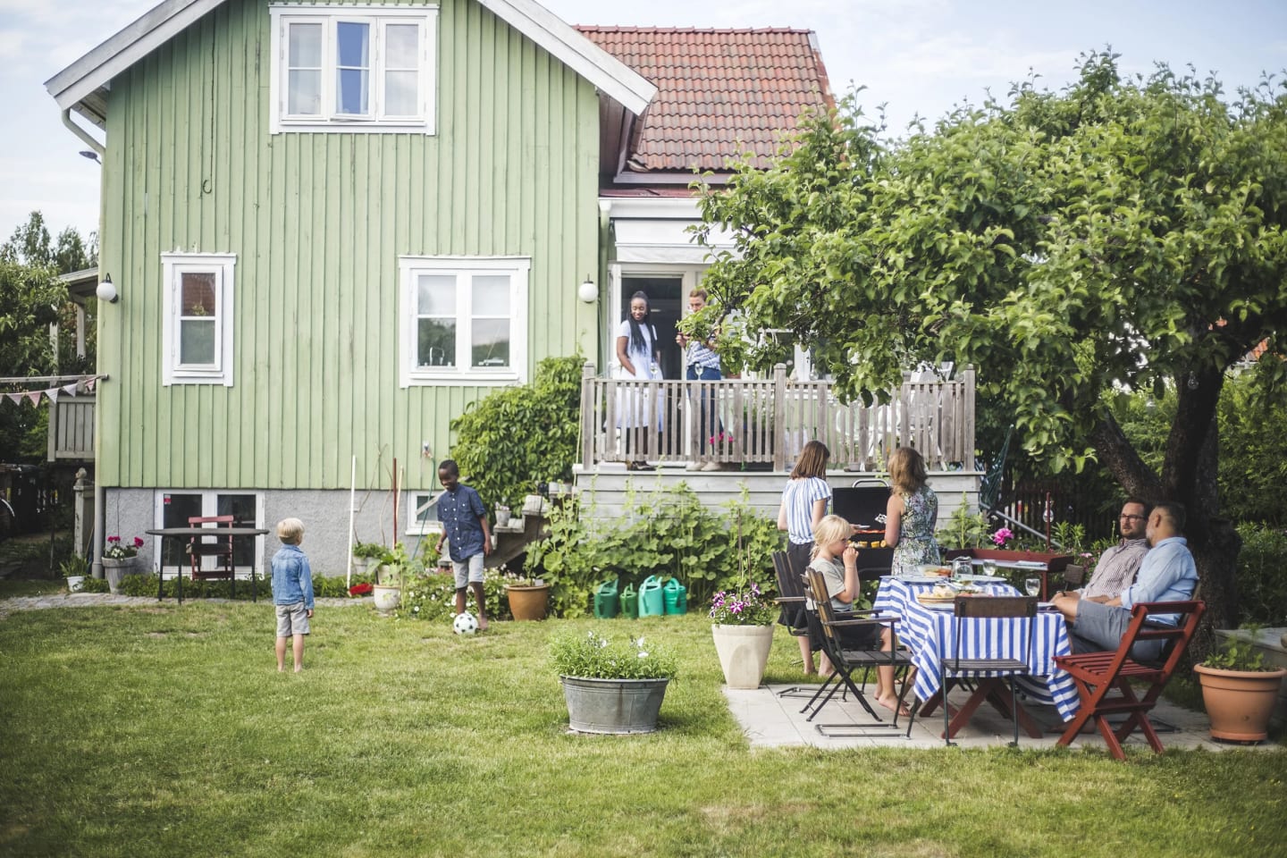 Flera folk i förgrunden sitter i trädgården och fikar. I bakgrunden är det ett grönt hus med fler folk på altanen.