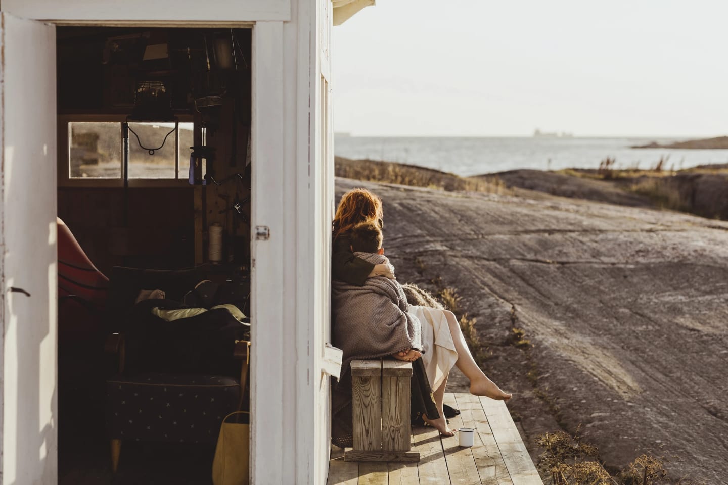 Mamma och son sitter på en bänk intill ett litet hus omringat av klippor och hav.