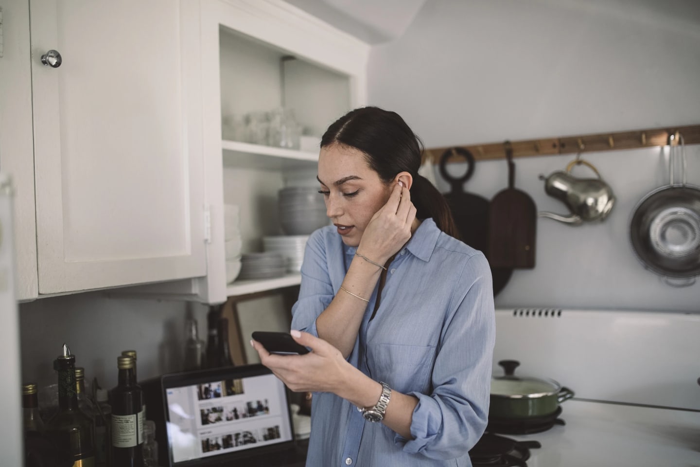 Kvinna står i köket och sätter i hörlurar och tittar ner i telefonen som för att ringa någon.