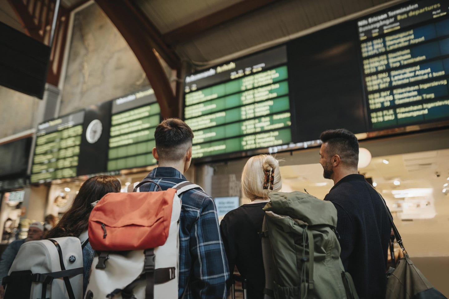 Ett gäng ungdomar står vid en terminal med ryggsäckar och tittar upp mot tavlan.