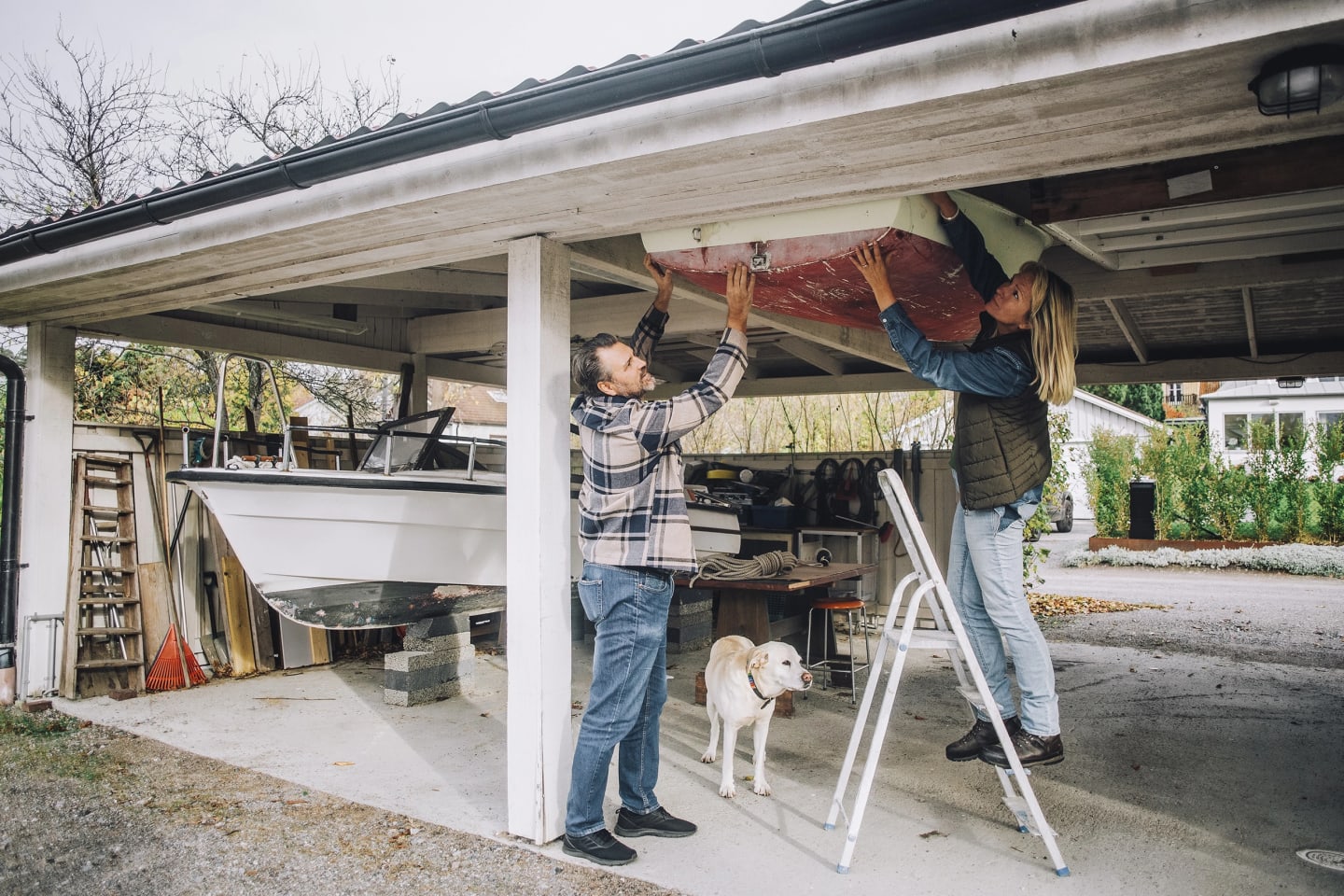 Man och kvinna fixar med båt under sin carport. Vid sidan står en hund och tittar.