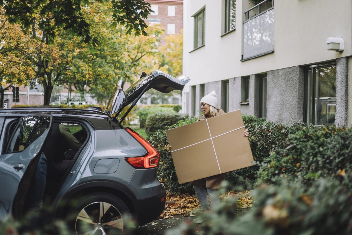 Kvinna packar in en stor kartong i baksätet på bil. Bilen stor precis utanför lägenhetsport.