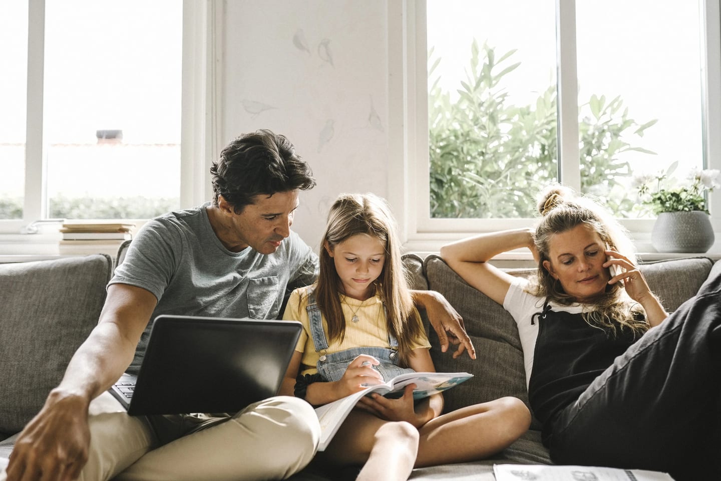 Bild på en familj som sitter i soffan. Mamman pratar i telefon och pappan och dottern tittar i en bok.