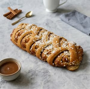 En gyllenbrun kanellängd med flätat eller klippt mönster, toppad med pärlsocker. Den ligger på ett fat och ser saftig ut med synlig kanelfyllning mellan lagren.