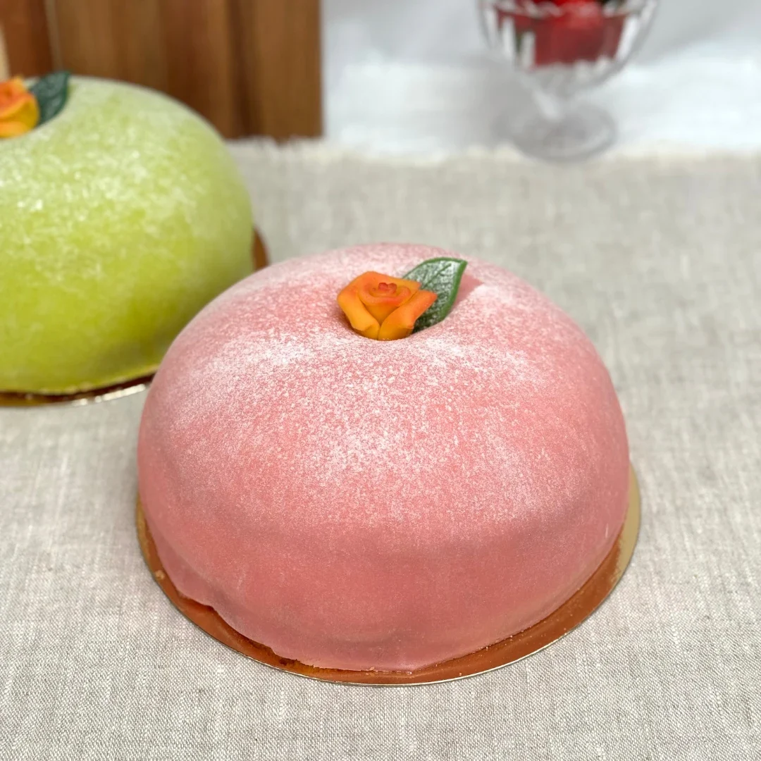 Prinsesstårta med rosa marsipan och en orange marsipanros på toppen, en grön prinsesstårta syns i bakgrunden.