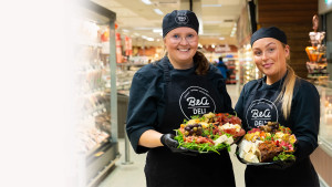 Två glada medarbetare som står framför delikatessen och håller i varsin charkbricka.